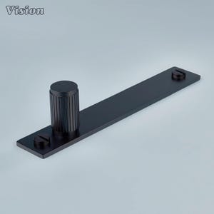 May include: A black metal door pull with a cylindrical handle and a rectangular base. The handle has vertical grooves, and the base has two screws. The word "Vision" is in the top left corner.