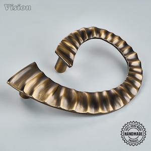 May include: A curved, antique brass-toned metal cabinet pull with a fluted, scalloped edge design. The hardware features a semi-circular shape with two mounting posts on the underside. The word Vision is in the top left corner of the image.