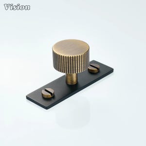May include: A bronze-colored cabinet knob with a cylindrical, ridged handle and a flat, rectangular black base. Two bronze screws secure the knob to the base. The word "Vision" is in the upper left corner.
