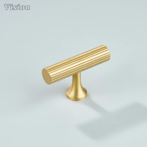 Puede incluir: Un tirador de armario de color dorado con un asa cilíndrica con ranuras verticales. El tirador está montado sobre una base corta y redondeada. La palabra "Vision" está en la esquina superior izquierda.