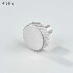 Può includere: Un pomello per armadio in argento spazzolato con una faccia circolare e un bordo strutturato a motivo di diamanti. Il pomello ha uno stelo cilindrico per il montaggio. La parola "Vision" è nell'angolo in alto a sinistra.