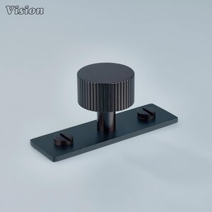 May include: A dark bronze cabinet pull with a cylindrical, ridged handle and a rectangular base. Two screws secure the base. The word "Vision" is in the upper left corner.