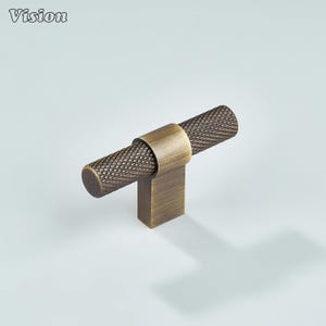 Puede incluir: Un tirador de armario de color bronce con un mango cilíndrico texturizado y una base cuadrada. El mango tiene un patrón de diamantes. La palabra "Vision" está en la esquina superior izquierda.