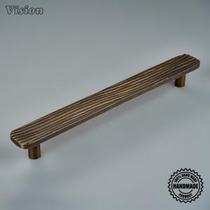 May include: A long, linear cabinet handle with a dark bronze finish and a textured, ribbed surface. The handle features two cylindrical mounting posts on the underside. The word Vision is printed in the top left, and a 100% handmade product seal is in the bottom right.
