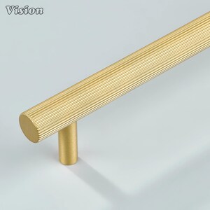 Pode incluir: Um puxador de armário dourado com uma pega cilíndrica ranhurada e uma base cilíndrica. A palavra "Vision" está impressa no canto superior esquerdo. O hardware é projetado para móveis e armários.