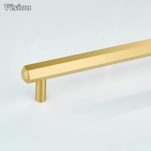 May include: A gold-colored cabinet pull with a geometric, hexagonal design. The pull has a cylindrical post and a long, flat bar with multiple facets. The word "Vision" is in the upper left corner.