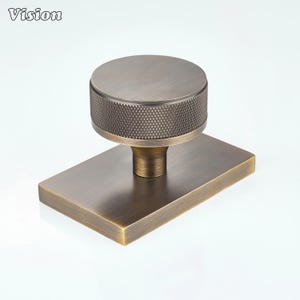 May include: A round cabinet knob with a textured, diamond-patterned knurled edge and a smooth top, attached to a rectangular base plate. The hardware features a brushed, antique bronze finish with visible metallic grain and dark undertones.