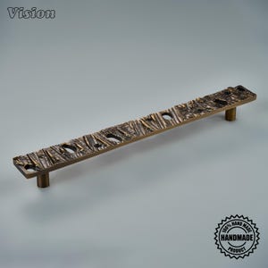 May include: A long, rectangular cabinet pull handle with a dark bronze finish and a textured, bark-like surface. The handle features a series of irregular, recessed openings along its length and is supported by two cylindrical mounting posts.