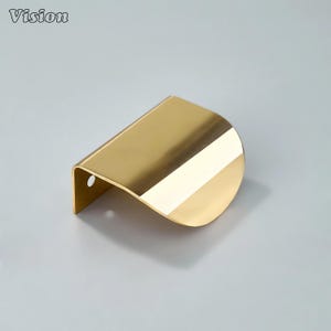 May include: A gold-colored cabinet pull with a curved, wave-like design. The pull has a smooth, reflective surface and a small hole on one end for mounting. The word "Vision" is in the upper left corner.