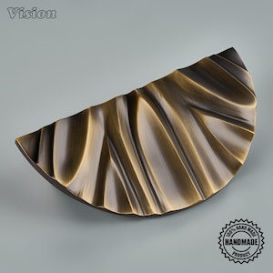 May include: A semi-circular cabinet pull with a brushed bronze finish and a textured, rippled surface. The hardware features a distinct wave-like pattern across its face. The word Vision is printed in the top left corner and a 100% handmade product seal.