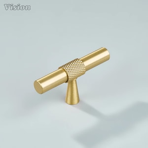 May include: A gold-colored cabinet pull with a T-bar design. The center section features a diamond-patterned texture, while the ends are smooth. The pull is mounted on a small, conical base. The word "Vision" is in the top left corner.