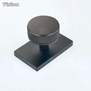 Pode incluir: Um puxador de armário preto fosco com um topo circular e uma borda serrilhada com padrão de diamante texturizado, montado em uma placa de base retangular. A palavra Vision está impressa no canto superior esquerdo da imagem.