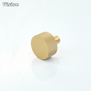 May include: A gold-colored cabinet knob with a textured, diamond-patterned grip. The knob has a cylindrical shape with a flat, circular face and a small, cylindrical post for attachment. The word "Vision" is in the upper left corner.