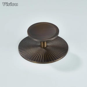 May include: Bronze cabinet knob with a circular top and a sunburst pattern on the base. The knob has a vintage look and is suitable for drawers or cabinets. The word "Vision" is in the top left corner.