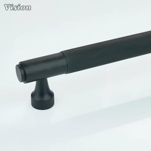 May include: A black cabinet pull with a textured, diamond-patterned grip. The pull has a cylindrical handle and a T-shaped base. The word "Vision" is in the upper left corner.