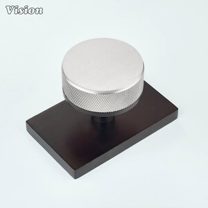 May include: A silver-coloured circular knob with a textured, knurled edge mounted on a rectangular, dark brown base. The top surface of the knob is smooth and flat, while the side features a cross-hatched pattern for grip. The word Vision is visible.