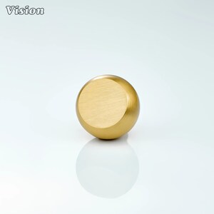 Puede incluir: Un pomo de armario esférico de color dorado, con una cara plana y circular. El pomo tiene un acabado liso y pulido y está sobre un fondo blanco. La palabra "Vision" está en la esquina superior izquierda.