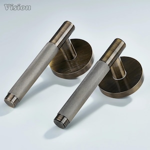 May include: Two antique bronze door handles with a textured grip and circular backplates. The handles are cylindrical with a diamond pattern. The Vision logo is in the top left corner.