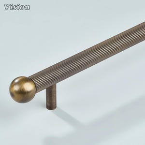 Pode incluir: Puxador de armário em tom bronze com barra canelada e extremidade esférica. O puxador possui um poste cilíndrico para montagem. A palavra "Vision" está no canto superior esquerdo. Adequado para armários e gavetas.
