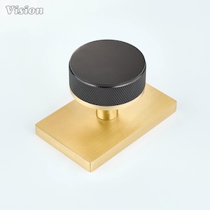 May include: A cabinet knob featuring a circular black top with a textured knurled edge, mounted on a rectangular brushed gold base. The knob has a smooth, flat black surface on top and a metallic gold stem connecting the knob to the base plate.