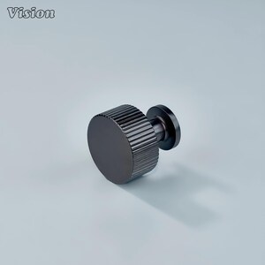 May include: A dark grey cabinet knob with a ridged, circular handle and a smaller, flat base. The word "Vision" is in the upper left corner. The knob is set against a light grey background.