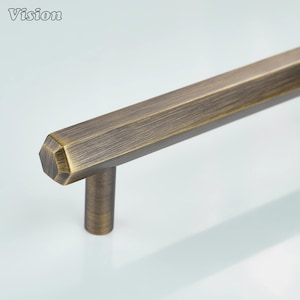 Puede incluir: Primer plano de un tirador de armario de color bronce con un diseño hexagonal. El tirador tiene un asa cilíndrica y una barra larga y geométrica. La palabra "Vision" está en la esquina superior izquierda.
