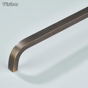 May include: A bronze-toned, curved cabinet pull with a rectangular bar and a rounded corner. The Vision logo is in the upper left corner. The hardware has a brushed finish and is set against a light background.
