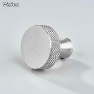 May include: A brushed silver cabinet knob with a circular face and a textured, diamond-patterned edge. The knob has a cylindrical stem and a smooth, flat surface. The word "Vision" is in the top left corner.