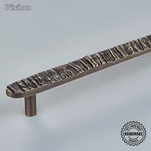 May include: A long, horizontal metal cabinet pull handle with a dark bronze finish. The surface features a rugged, textured pattern of vertical ridges and grooves. A cylindrical mounting post is visible on the left side. The word Vision is in the top left corner, and a circular 100% handmade product stamp is in the bottom right.