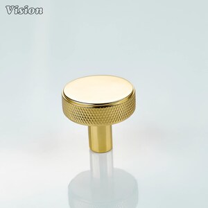 Op de afbeelding: Een goudkleurige kastknop met een getextureerde, diamantpatroon grip en een gladde, cirkelvormige bovenkant. De knop heeft een cilindrische basis en staat tegen een witte achtergrond. Het woord "Vision" staat in de linkerbovenhoek.