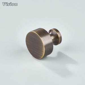 May include: A bronze-colored cabinet knob with a circular face and a smaller, cylindrical base. The knob has a brushed metal finish and is designed for furniture. The word "Vision" is in the top left corner.