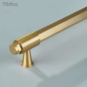 Könnte beinhalten: Ein gebürsteter goldfarbener Möbelgriff mit einer langen sechseckigen Stange, einer zylindrischen Endkappe und einem konisch zulaufenden Befestigungssockel. Der Metallbeschlag hat eine matte Oberfläche und ein klares, geometrisches Design.