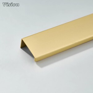 May include: A gold-colored cabinet pull with a modern, minimalist design. The pull is rectangular with a folded edge, creating a sleek, handle-less appearance. The word "Vision" is in the upper left corner.