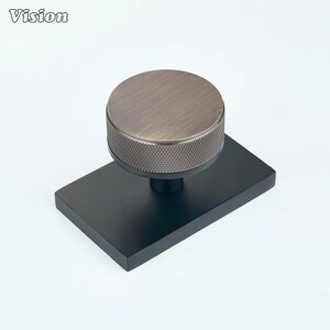 Pode incluir: Um puxador circular com topo em bronze escovado e borda lateral texturizada com padrão de diamante, montado sobre uma placa de base retangular preta fosca. A palavra Vision está impressa no canto superior esquerdo da imagem.
