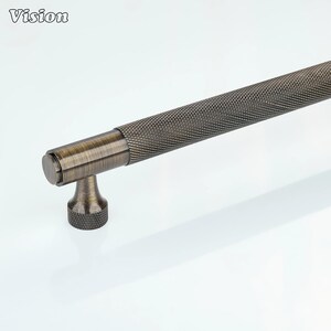 Könnte beinhalten: Ein bronzefarbener Schrankgriff mit einem strukturierten, rautenförmigen Griff. Der Griff hat ein zylindrisches Design mit einem kleinen, strukturierten Sockel und einer runden Endkappe. Das Wort "Vision" befindet sich in der oberen linken Ecke.