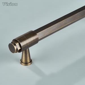 Könnte beinhalten: Ein langer, sechseckiger Möbelgriff mit gebürstetem Antik-Bronze-Finish. Der Beschlag verfügt über einen zylindrischen Befestigungspfosten, der mit einem ausgestellten, kreisförmigen Sockel verbunden ist. Das Wort Vision steht oben links.