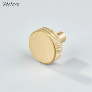 May include: A gold-colored cabinet knob with a textured, diamond-patterned grip and a smooth, circular face. The knob has a cylindrical stem for mounting. The word "Vision" is in the upper left corner.