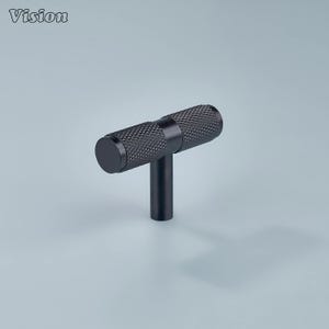 May include: A black T-bar cabinet pull with a textured, diamond-patterned grip. The pull has a cylindrical handle and a vertical post for mounting. The word "Vision" is in the upper left corner.