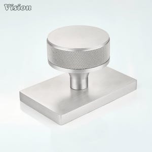 Puede incluir: Un pomo de metal plateado para muebles con una parte superior circular con un borde moleteado de patrón de diamante, montado sobre una placa base rectangular con acabado cepillado. La palabra Vision está impresa en la esquina superior izquierda.