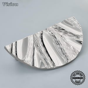 Può includere: Una maniglia per mobili argentata a forma di semicerchio con una finitura lucida altamente riflettente. La superficie presenta un motivo organico strutturato di creste e scanalature in rilievo che imitano le venature del legno o l'acqua.