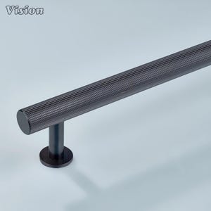 May include: A dark gray, textured metal bar with a cylindrical design, supported by a vertical post and a circular base. The word "Vision" is in the upper left corner. This hardware is likely for a door or cabinet.