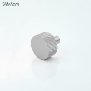 May include: A silver cabinet knob with a textured grip and a cylindrical shape. The knob has a flat, circular face and a smaller, cylindrical post for mounting. The word "Vision" is in the upper left corner.