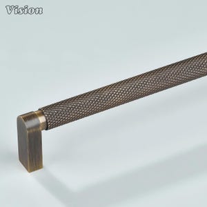 May include: Bronze-toned cabinet pull with a textured, diamond-patterned handle and a square end. The word "Vision" is in the upper left corner. This hardware is suitable for drawers and cabinets.