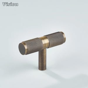 May include: A bronze-toned cabinet pull with a textured, diamond-patterned handle. The T-shaped pull features a cylindrical handle with a central band and a short, cylindrical stem. The word "Vision" is in the upper left corner.