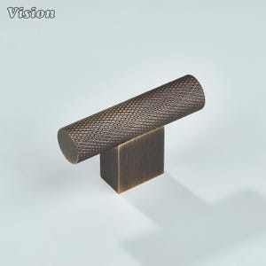 May include: Bronze cabinet pull with a textured, cylindrical handle and a square base. The handle has a diamond pattern. The word "Vision" is in the upper left corner. This hardware is suitable for drawers and cabinets.