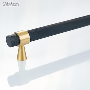 Peut inclure: Une poignée de meuble composée d'une longue barre cylindrique à la texture noire mate moletée. La poignée est soutenue par une base conique dorée brossée et un anneau de raccordement doré assorti. Le mot Vision est inscrit en haut à gauche.