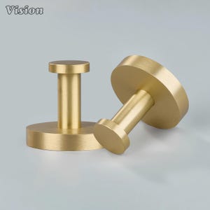 May include: Two brass-colored wall hooks. Each hook has a circular base and a cylindrical post topped with a smaller circular head. The hooks are made of metal and have a matte finish. The word "Vision" is in the top left corner.