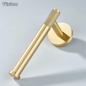 May include: A gold-colored, modern toilet paper holder. The holder features a cylindrical bar with a textured grip and a circular wall mount. The word "Vision" is in the top left corner.