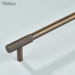 May include: Bronze-toned cabinet pull with a textured, diamond-patterned grip. The Vision logo is in the upper left corner. The handle has a cylindrical shape and a vertical post for mounting. Ideal for kitchen or bathroom cabinetry.