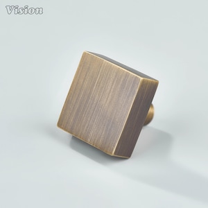 Pode incluir: Um puxador de armário quadrado com acabamento em bronze escovado. O puxador tem um design geométrico simples e uma pequena base redonda. A palavra "Vision" está no canto superior esquerdo.